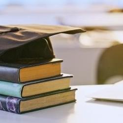 Three hardback books stacked on a desk with a graduate mortarboard on top, a silver laptop sits on its left.