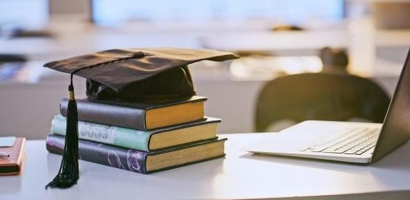 Three hardback books stacked on a desk with a graduate mortarboard on top, a silver laptop sits on its left.